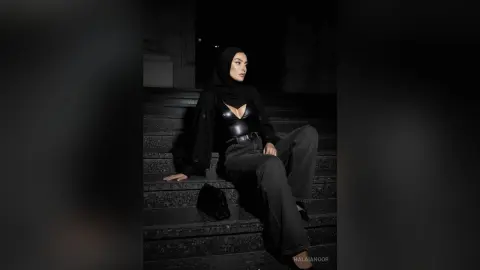 Elegant woman in a black hijab and chic dark outfit sitting on stone stairs at night.