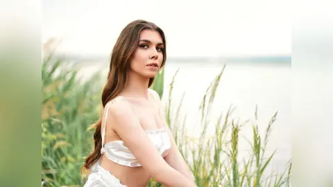 Young woman in a white summer camisole relaxing by a serene lakeside with tall green reeds.