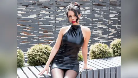 Elegant woman in a sleek black satin halter dress posing against a modern textured wall in a garden setting.