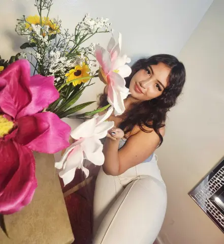 Smiling young woman in white pants sitting by vibrant pink and yellow flowers in a bright indoor setting.