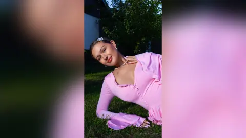 Smiling woman in a pink long-sleeve blouse relaxing outdoors in a sunny garden setting.