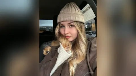 LillyEyes wears a tan beanie and a brown coat while sitting in a car, showing off her long blonde hair and light eyes.