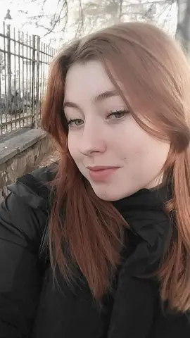 Smiling young woman with long red hair wearing a black winter jacket in a soft outdoor setting.