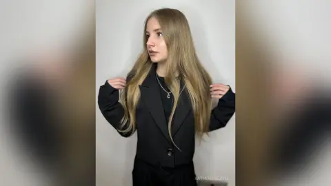 Blonde woman in a chic black blazer and silver necklace posing against a white background with a minimalist mood.