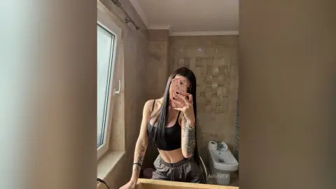 AriaVex takes a mirror selfie wearing a black tank top and grey shorts, showing her long dark hair and arm tattoos.
