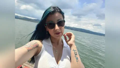 Woman with blue hair and sunglasses smiling on a sunny boat trip over a calm lake.