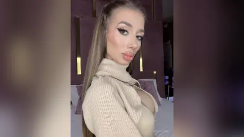 DeviNessa poses in a tan turtleneck sweater that shows off her cleavage, looking over her shoulder with long blonde hair.