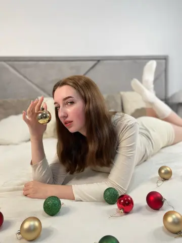 Young woman in a cream long-sleeve dress lying on a bed surrounded by colorful Christmas ornaments.