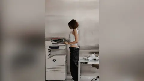 Woman in white tank top and black jeans standing by a printer in a minimalist office setting.