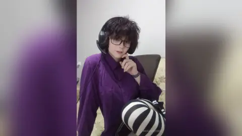 Young woman with glasses and headphones wearing a purple button-down shirt in a cozy indoor setting.
