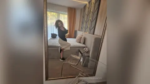 Woman in black top and white trousers taking a mirror selfie in a modern, cozy bedroom.