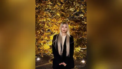LurlineLacy poses in a black blazer with long blonde hair against a background of golden autumn leaves.