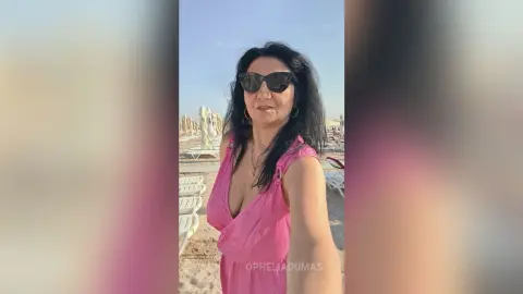 Smiling woman in a pink summer dress and sunglasses enjoying a sunny day at a sandy beach resort.