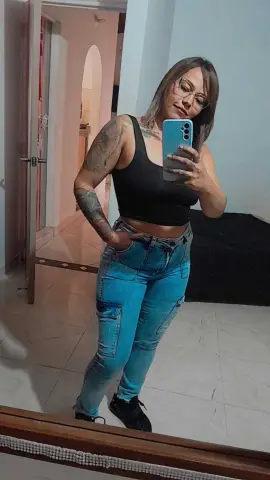Tattooed woman in a black crop top and blue jeans taking a confident mirror selfie in a modern room.