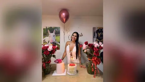 Smiling woman in a white dress celebrating with red roses, cake, and a pink balloon in a festive indoor setting.