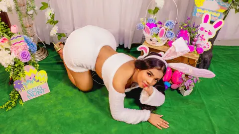 Sra_Canelita poses on all fours in a white fuzzy bunny outfit and ears, surrounded by colorful Easter decorations and flowers.