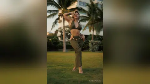Stylish woman in olive green bikini top and trousers posing in a tropical garden at sunset.
