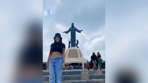 Woman in black crop top and jeans standing before the Christ the Redeemer statue under a cloudy sky.