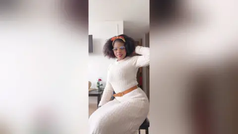 Smiling Black woman in a white knit sweater and headband posing in a bright, minimalist living room.