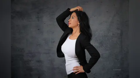 Elegant woman in a black blazer and white top posing confidently against a textured gray studio background.