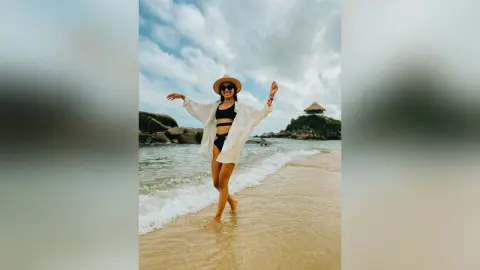 Happy woman in a white cover-up and sun hat walking on a tropical beach under a cloudy sky.