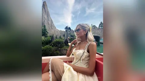 NickyAshcroft poses outdoors in a light yellow dress and sunglasses, sitting on a red seat with her blonde hair styled down.