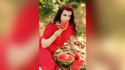 Young woman in a red polka dot dress eating watermelon during a sunny summer picnic outdoors.