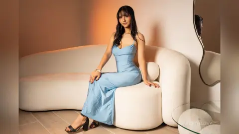 Elegant woman in a light blue slip dress sitting on a white curved sofa in a warm, minimalist room.