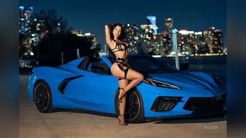 Alison poses against a blue sports car wearing black strappy lingerie and high heels against a night city skyline.