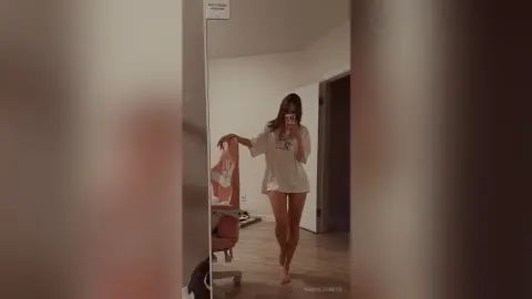 TarenLeonetti stands in a room wearing a white t-shirt, posing with one hand on a pink chair and her legs crossed.