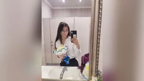 Young woman in a white blouse holding a blue flower bouquet during a mirror selfie in a bright bathroom.
