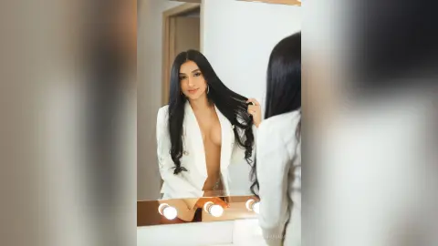 AlessandraSanit poses in a white blazer, running her hands through her long black hair while looking into a lit mirror.
