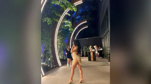 NicoleBella stands in a lit outdoor garden setting wearing a short, gold-colored dress that exposes her back and legs.