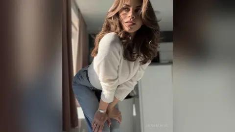 AnaMathers leans forward wearing a white long-sleeve top and blue jeans, letting her long brown hair fall over her shoulders.