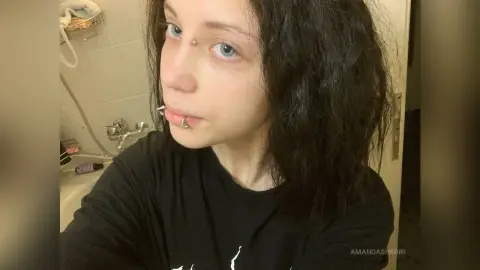 Young woman with curly dark hair and facial piercings wearing a black t-shirt in a bright bathroom setting.