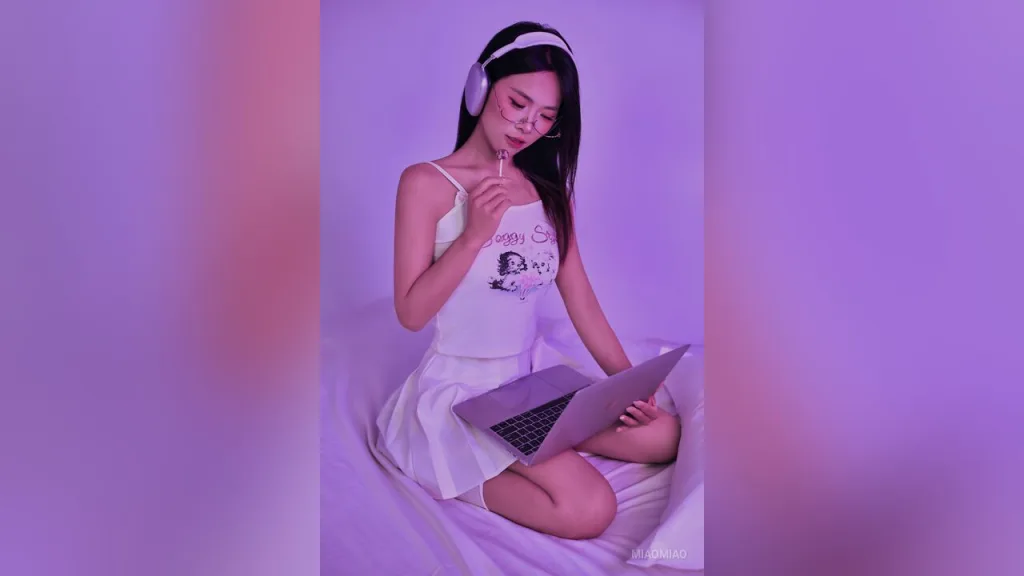 miaomiao — miaomiao sits on a bed wearing white loungewear and headphones while looking at a laptop.