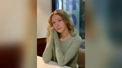 Blonde woman in a sage green long-sleeve shirt posing with a thoughtful expression in a modern indoor setting.