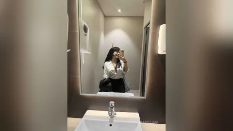 Young woman in a white blouse taking a mirror selfie in a modern minimalist bathroom.