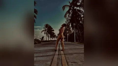 JulianaNavarro poses naked on a road lined with palm trees, showing her bare back and buttocks while looking back over her shoulder.