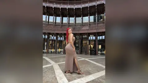Stylish woman in a long beige slit dress posing confidently in a modern luxury shopping plaza.