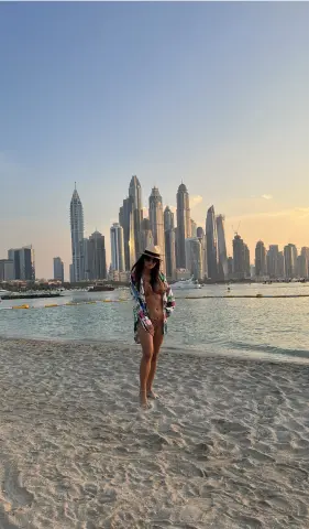 Woman in a patterned shirt and bikini standing on a sandy beach with a Dubai skyline sunset background.
