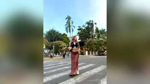 Stylish woman in a black bikini top and red patterned maxi skirt walking across a street in a tropical setting.