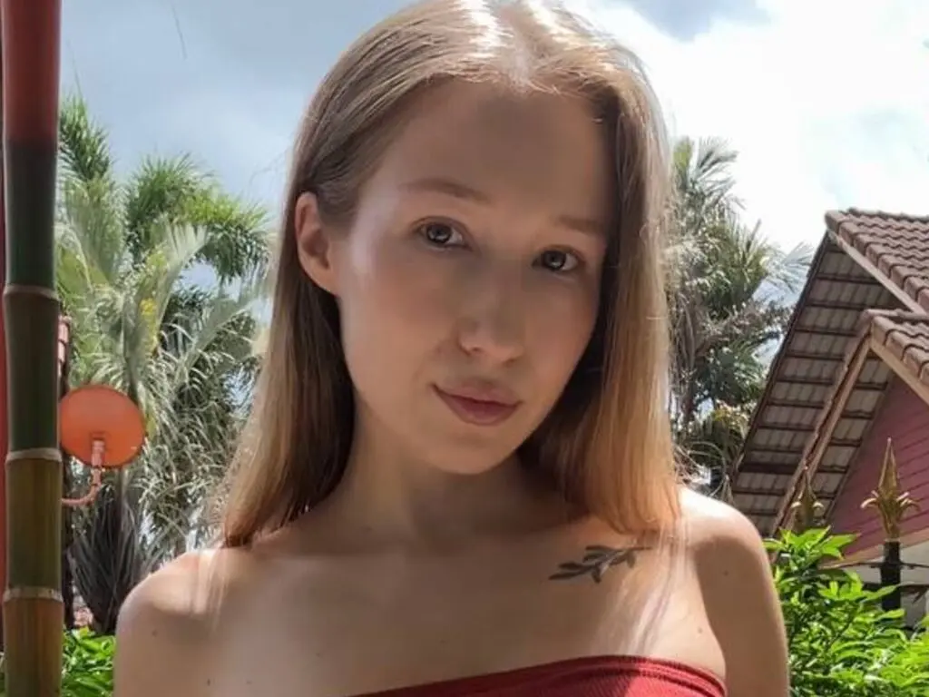 Young woman with long blonde hair in a red strapless top posing outdoors near tropical palm trees.