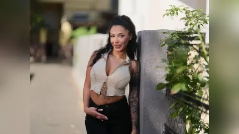 Smiling woman in a beige cropped vest and black jeans leaning against a wall in a sunny outdoor setting.
