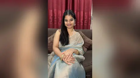 Sejuti sits on a brown couch wearing a light blue saree and a white pearl necklace, looking at the camera with long dark hair.