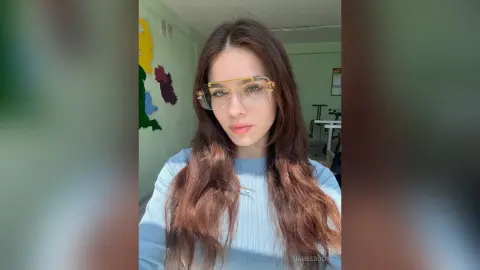 Young woman with long brown hair wearing clear glasses and a light blue ribbed sweater in a bright room.