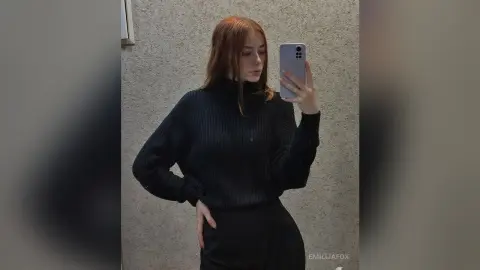 Redheaded woman in a cozy black ribbed turtleneck sweater taking a mirror selfie in a minimalist room.