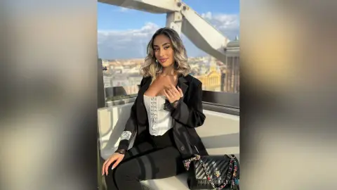 Stylish woman in a black blazer and white corset sitting on a rooftop terrace under a bright blue sky.