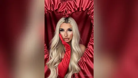 Glamorous blonde woman in red satin gloves posing against a romantic red heart-shaped fabric backdrop.