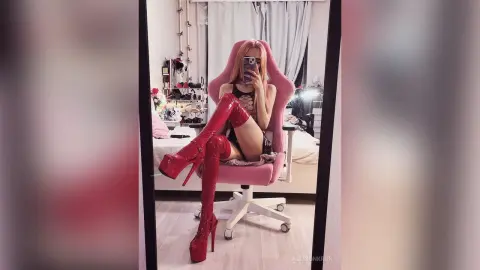 AllisonKron sits in a pink chair wearing black lace lingerie and red patent leather thigh-high boots while taking a mirror selfie.